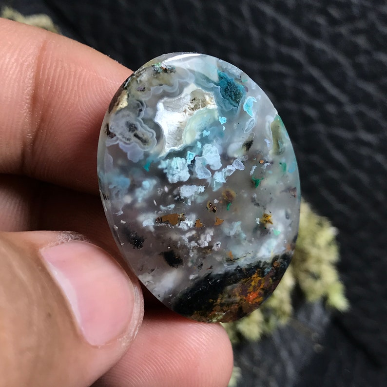May include: A cabochon cut gemstone with a cloudy, white, and blue agate pattern. The stone has a dark grey, black, and brown base with flecks of gold and green.