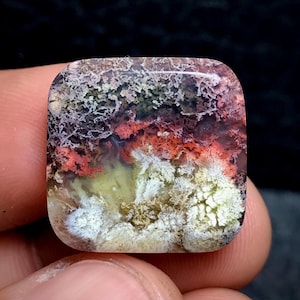 Könnte beinhalten: Ein quadratischer Edelstein mit einem einzigartigen, natürlichen Muster. Der Stein weist eine Mischung aus Farben auf, darunter Weiß, Rot, Grün und Braun, wodurch ein strukturiertes, organisches Aussehen entsteht. Der Edelstein wird zwischen Fingern gehalten, was seine komplizierten Details zeigt.