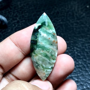 Può includere: Un cabochon di agata verde, bianco e marrone con forma ovale appuntita. La pietra presenta un motivo a spirale ed è traslucida.