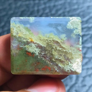 Peut inclure: Un cabochon d'agate vert, blanc et orange avec un motif ressemblant à un paysage. La pierre est translucide et a une surface lisse et polie.