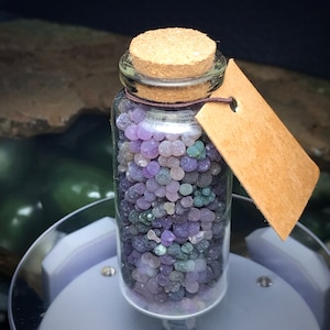 Puede incluir: Un pequeño frasco de vidrio lleno de piedras de ágata de color púrpura, verde y blanco. El frasco tiene una tapa de corcho y una etiqueta de papel marrón.