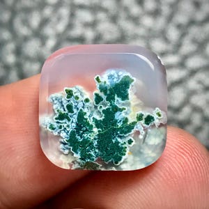 May include: A square-shaped, polished gemstone with a translucent appearance. The stone features a vibrant green and white moss-like pattern within, set against a pale pink and clear background. The edges are rounded, giving it a smooth, tactile feel.