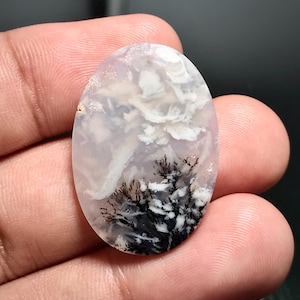 Puede incluir: Un cabujón de ágata blanco y gris con inclusiones dendríticas negras. La piedra tiene un patrón nublado y arremolinado.