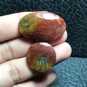 Peut inclure: Deux cabochons d'agate, l'un ovale et l'autre rond, avec un motif de couleur rouge, orange et vert. Les pierres ont un aspect naturel et organique.