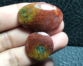 Pode incluir: Dois cabochões de ágata, um oval e outro redondo, com um padrão de cor vermelho, laranja e verde. As pedras têm um aspecto natural e orgânico.
