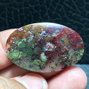 Pode incluir: Um cabochão de ágata verde, vermelho e marrom com uma superfície lisa e polida. A pedra tem um padrão único e espiralado de cores e texturas.