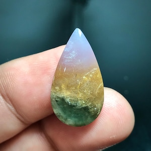 May include: A teardrop-shaped gemstone with a gradient of colors. The top is a pale lavender, transitioning to a golden-brown center, and ending with a green base. The stone has a polished, smooth surface.