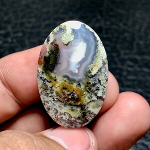 Könnte beinhalten: Ein glatter, ovaler Achat-Cabochon mit einem weißen, grauen und braunen Muster. Der Stein hat eine einzigartige, moosartige Textur und eine Vielzahl von Farben, darunter Grün, Gelb und Rot.