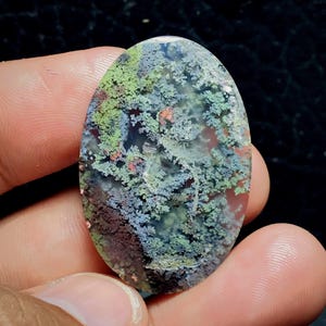 May include: An oval moss agate gemstone with a detailed, natural pattern. The stone displays a blend of green, blue, red, and brown colours, reminiscent of a small landscape. The polished surface accentuates the unique, organic design.