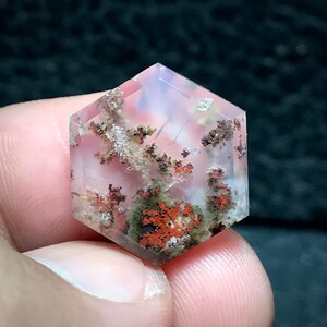 May include: A hexagonal clear quartz crystal with inclusions of red, brown, and green dendrites. The crystal is translucent and has a pink hue.