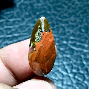 Może przedstawiać: Kabochoński kamień w kształcie łzy z jaspisu czerwonego, pomarańczowego i brązowego z białymi i szarymi wtrąceniami. Kamień ma polerowaną powierzchnię i jest naturalnym kamieniem szlachetnym.