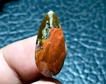 May include: A teardrop-shaped cabochon of red, orange, and brown jasper with white and grey inclusions. The stone has a polished surface and is a natural gemstone.
