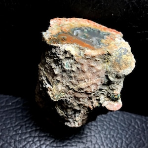 Puede incluir: Espécimen de piedra natural con una superficie rugosa y texturizada. La sección transversal revela una cavidad central. La piedra presenta una mezcla de colores, incluyendo beige, naranja, verde y blanco, sobre un fondo oscuro. La forma es irregular.