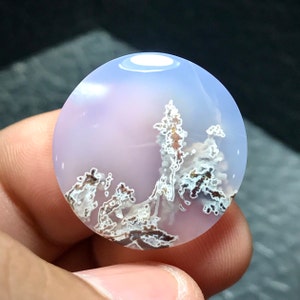 May include: A round, polished agate cabochon with a light blue and pink background. The stone features a dendritic pattern of brown and white inclusions, resembling a landscape.