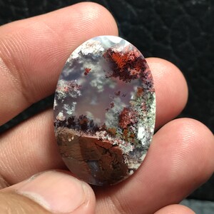 May include: A close-up of a polished oval-shaped agate gemstone with a white, gray, and brown base with red and green inclusions.