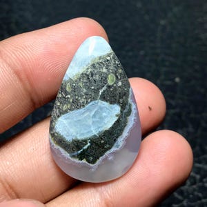 May include: A teardrop-shaped cabochon gemstone featuring a smooth, polished surface with a central patch of dark, mottled green and black matrix surrounded by translucent, milky white and pale lavender-grey agate banding.