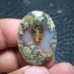 May include: An oval-shaped moss agate gemstone featuring a translucent, milky white base with distinct, intricate inclusions of deep green, olive, and rusty orange mineral deposits that resemble organic, moss-like patterns throughout the stone.