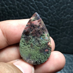 Puede incluir: Una gema en forma de lágrima con un patrón único. La piedra presenta una mezcla de tonos verdes, marrones y rojos, que recuerdan a un paisaje en miniatura. La superficie pulida resalta las texturas y colores naturales, creando un efecto visual llamativo.