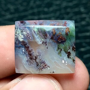 May include: A square-cut gemstone with a unique, natural pattern. The stone features a blend of colors, including white, green, brown, and blue, creating a landscape-like appearance. The polished surface reflects light, highlighting the intricate details.