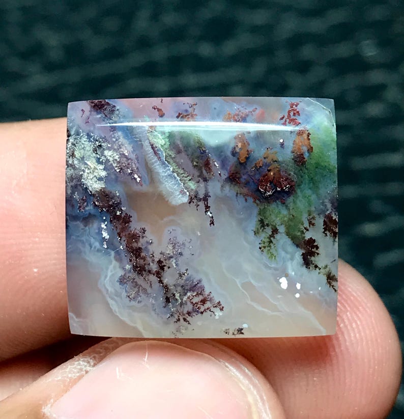 May include: A square-cut gemstone with a unique, natural pattern. The stone features a blend of colors, including white, green, brown, and blue, creating a landscape-like appearance. The polished surface reflects light, highlighting the intricate details.