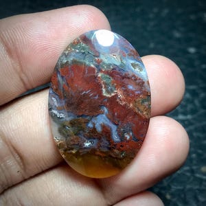 May include: A smooth, oval-shaped agate cabochon with a swirling pattern of red, brown, white, and gray. The stone has a polished finish and is translucent.