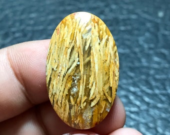 Puede incluir: Una piedra preciosa cabujón de color marrón claro translúcido con un patrón único de finas líneas marrón claro. La piedra preciosa tiene forma ovalada y una superficie lisa y pulida.