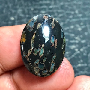 May include: Oval-shaped gemstone with a polished, glossy surface. The stone is primarily black, with irregular patterns of blue, green, and brown inclusions. The gemstone is held between fingers.