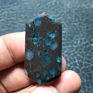 May include: A polished, hexagonal-shaped gemstone cabochon featuring a dark brown, nearly black matrix base with embedded, translucent, circular and irregular inclusions of vibrant teal and light blue mineral deposits throughout the surface.