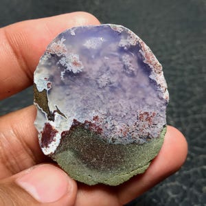 May include: A polished, oval-shaped gemstone with a layered appearance. The top section is a translucent lavender color, transitioning to white and red patterns, and ending with a dark green base. The stone is held in a hand.
