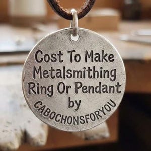 May include: A round, silver-toned metal pendant with a textured, weathered finish. The pendant features black engraved text that reads: Cost To Make Metalsmithing Ring Or Pendant by CABOCHONSFORYOU. It hangs from a dark brown leather cord.