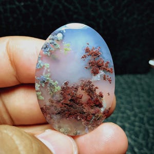 May include: An oval-shaped moss agate gemstone featuring a translucent, milky white base with distinct, branching dendrite inclusions in shades of deep rust red, muted green, and dark gray, creating a natural, tree-like pattern within the stone.