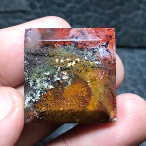 Könnte beinhalten: Ein quadratischer, mehrfarbiger Achat-Cabochon mit polierter Oberfläche. Der Stein zeigt eine Vielzahl von Farben, darunter Rot, Orange, Braun, Grün und Weiß. Der Achat hat ein einzigartiges, wirbelndes Muster.
