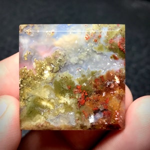 May include: A polished agate cabochon with a unique landscape pattern. The stone features shades of green, brown, white, and red, creating a natural abstract design.