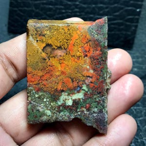 May include: A multi-coloured, roughly cut stone with a vibrant mix of hues. The stone features a blend of orange, yellow, red, green, and purple. The natural stone has a textured surface and irregular edges, showcasing its raw, unpolished state.