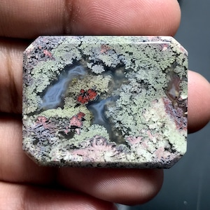 May include: A rectangular gemstone with a unique pattern of green, red, and gray hues. The stone's surface features a moss-like texture with intricate details, creating a natural and organic appearance. The gemstone is held between fingers.