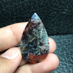 Könnte beinhalten: Ein tropfenförmiger Cabochon aus Achat mit einer Mischung aus schwarzen, weißen, braunen und roten Farben. Der Stein hat ein geflecktes, wolkiges Aussehen.