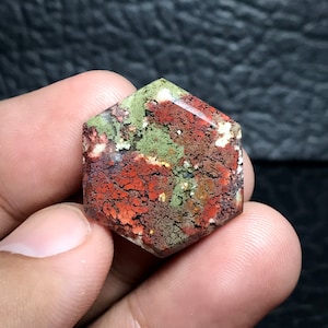 Pode incluir: Uma pedra preciosa cabochão hexagonal com um padrão vermelho, verde e branco. A pedra tem uma superfície áspera e texturizada.