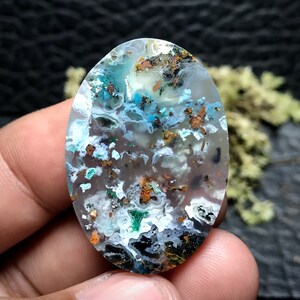 May include: A translucent oval cabochon gemstone with a white, blue, and green agate pattern. The stone is speckled with small, metallic copper inclusions.