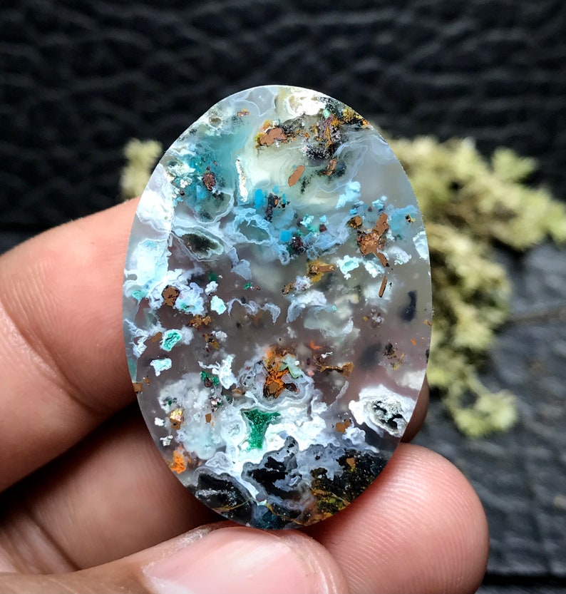 May include: A translucent oval cabochon gemstone with a white, blue, and green agate pattern. The stone is speckled with small, metallic copper inclusions.