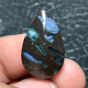 May include: A teardrop-shaped gemstone with a dark brown base, featuring irregular patterns of blue and teal. The polished surface reflects light, highlighting the stone's unique colour variations and natural textures. This gemstone is ideal for jewellery making.
