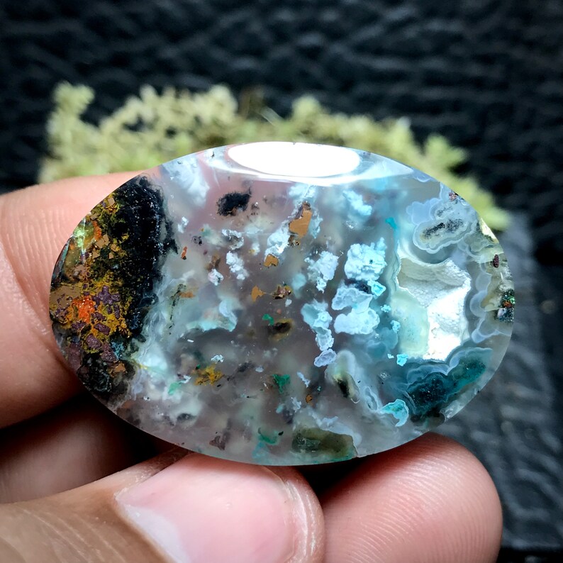 May include: A light blue, oval-shaped agate cabochon with a variety of inclusions, including black, brown, and gold flecks. The agate has a translucent appearance and shows a variety of patterns and textures.