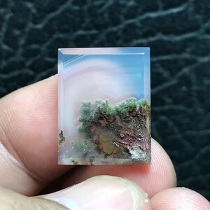 May include: A translucent, light pink agate cabochon with a landscape-like pattern of green, brown, and white inclusions. The agate is cut into a rectangular shape.