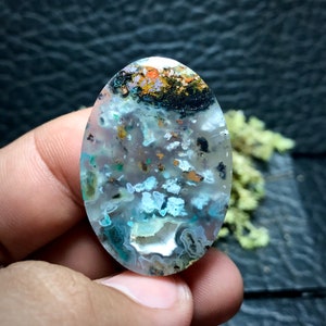 May include: A cabochon gemstone with a unique pattern of white, blue, green, and brown. The stone is oval shaped and has a smooth, polished surface.