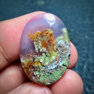 Peut inclure: Une pierre précieuse ovale avec un fond lavande translucide. La pierre présente un paysage d'inclusions vert mousse, marron et orange, ressemblant à une scène naturelle miniature. La surface polie reflète la lumière, soulignant les détails complexes.