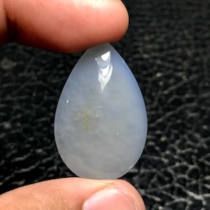 May include: A pear-shaped, pale blue gemstone with a polished surface. The stone has a subtle, cloudy appearance and a slight yellow inclusion. The gemstone is held between two fingers, showcasing its shape and color.