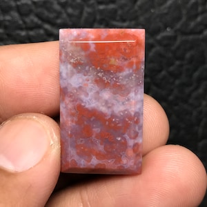 Puede incluir: Un cabujón rectangular de ágata rojo y blanco pulido con una superficie lisa y pulida. La piedra tiene un patrón de remolino de rojo, blanco y marrón.