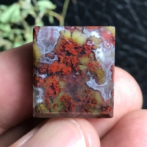 May include: A square-cut gemstone with a unique pattern of red, yellow, and white inclusions. The stone has a polished surface and a translucent appearance, revealing intricate internal details. The overall effect is a blend of earthy tones and natural beauty.