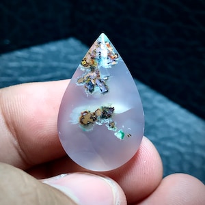 Peut inclure: Un cabochon d'agate en forme de larme avec une base translucide, violet clair. La pierre présente des inclusions de cuivre, d'oxyde de fer et de malachite verte.