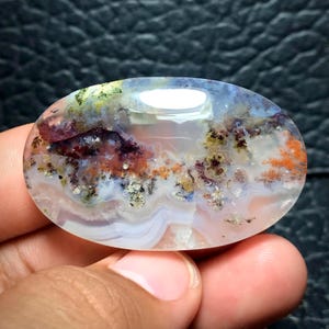 Op de afbeelding: Ovale, gepolijste edelsteen. De steen heeft een mix van kleuren, waaronder wit, oranje, paars en groen, waardoor een uniek, natuurlijk patroon ontstaat. De edelsteen wordt in een hand gehouden, wat de grootte en details benadrukt.