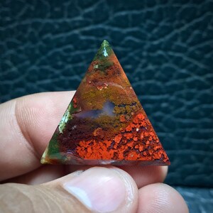Puede incluir: Una piedra de ágata pulida de forma triangular con una mezcla de colores rojo, naranja, marrón y verde. La piedra tiene un patrón único y moteado.